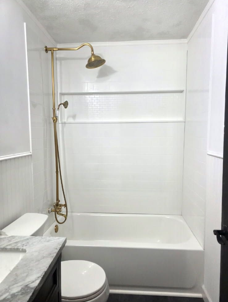 White bathroom with shower-tub, brass showerhead, toilet, and marble sink countertop.