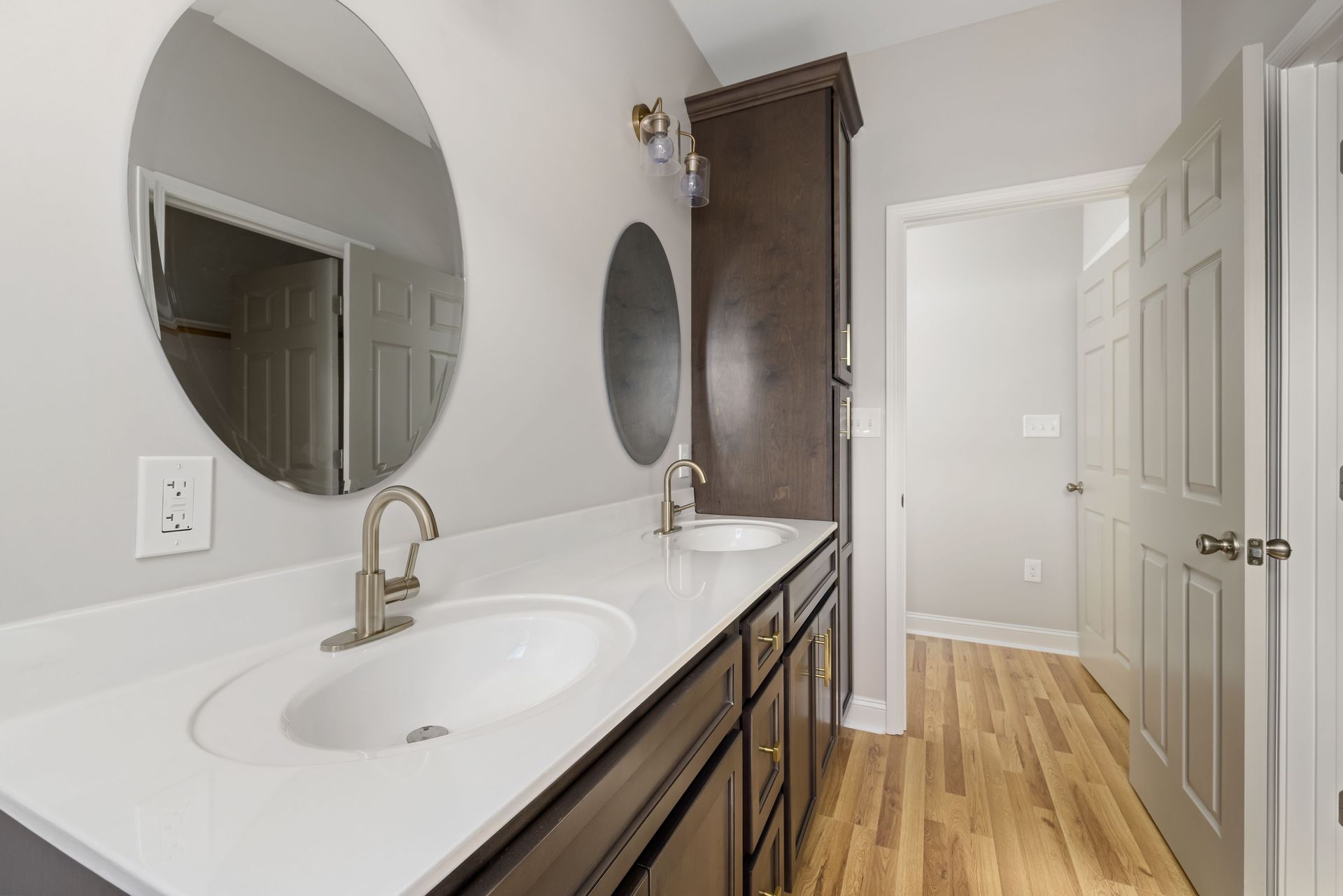 Bright bathroom vanity with round mirror, white countertop, and wood floor beside a shower doorway.
