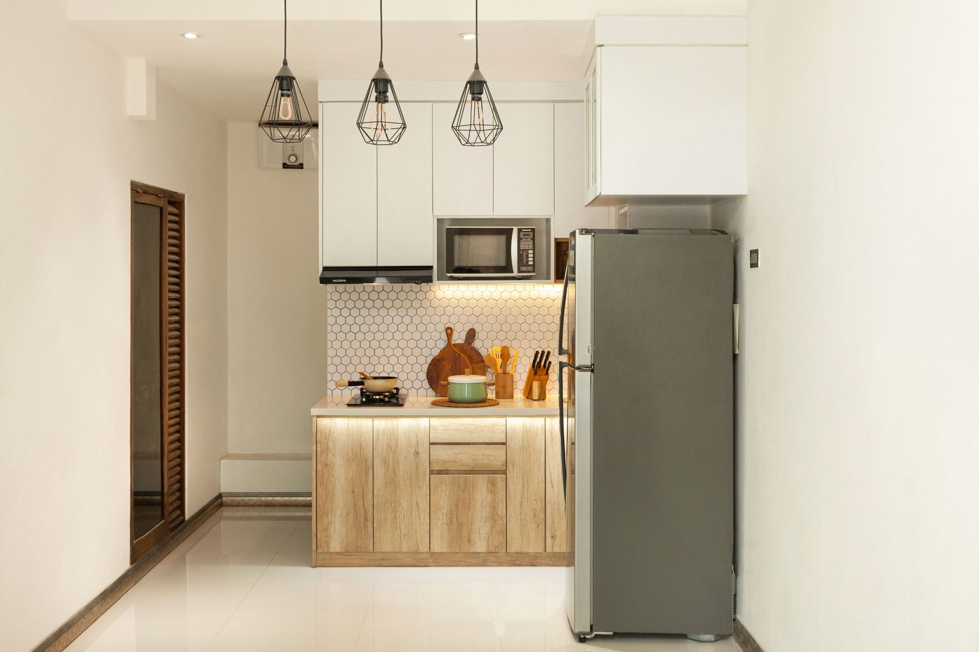 Small modern kitchen with wood cabinets, white walls, pendant lights, and a stainless-steel refrigerator
