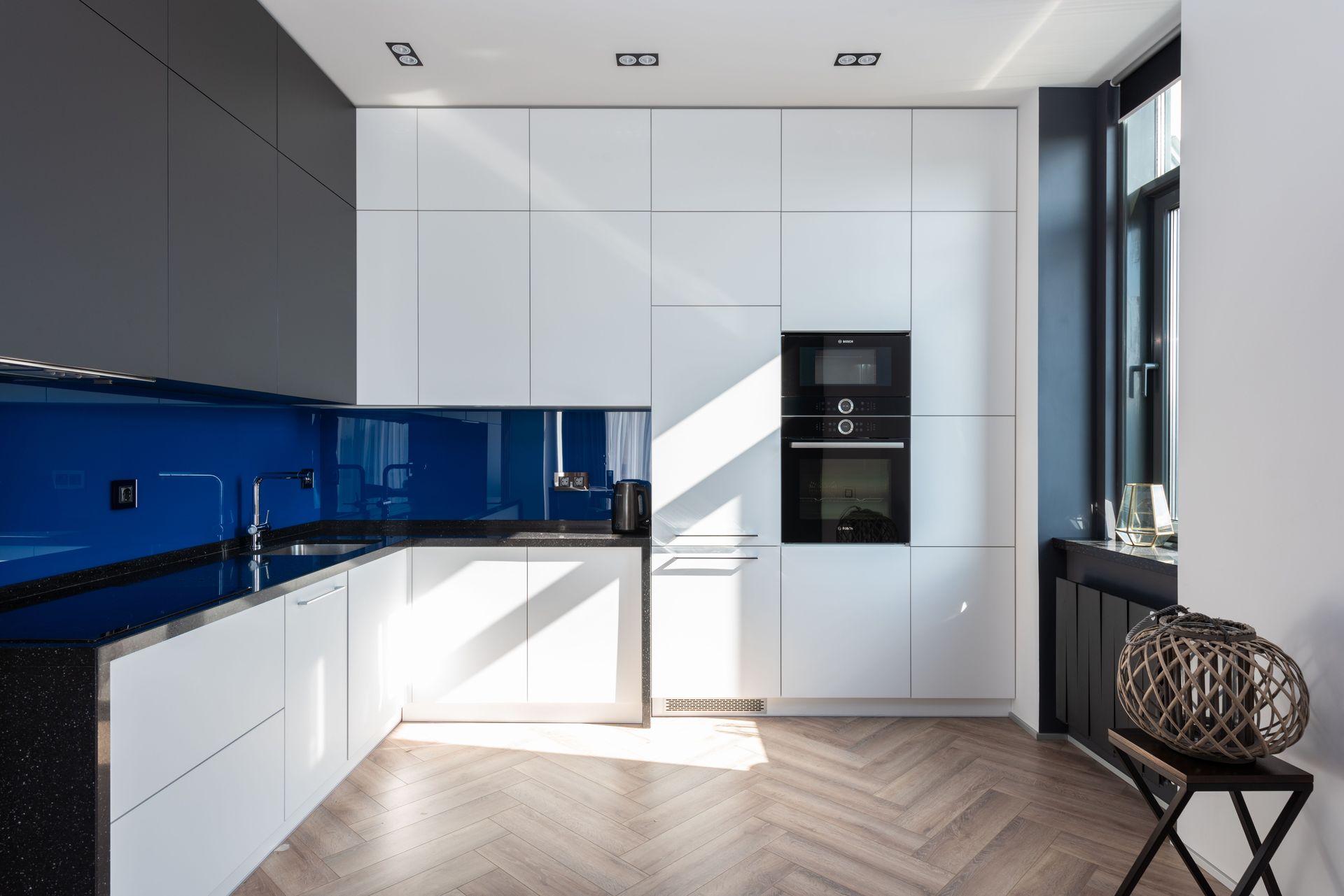 Modern white kitchen with blue backsplash, built-in oven, and herringbone wood floor.