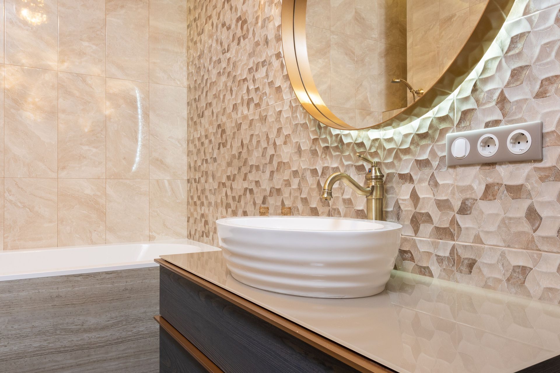 Modern bathroom vanity with white vessel sink, round mirror, beige tiled wall, and gray countertop.