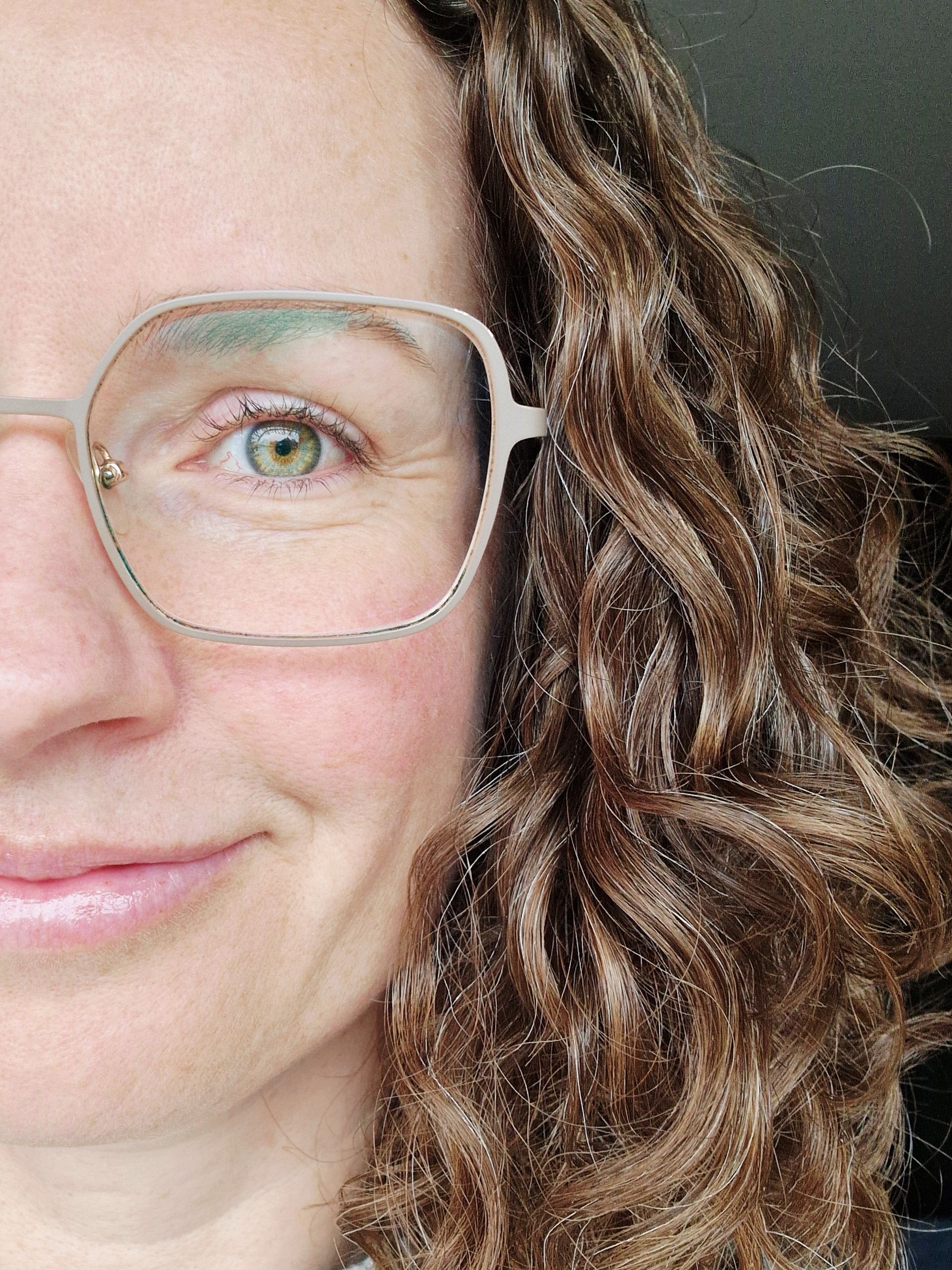 Een close-up van het gezicht van een persoon met een bril, golvend bruin haar.