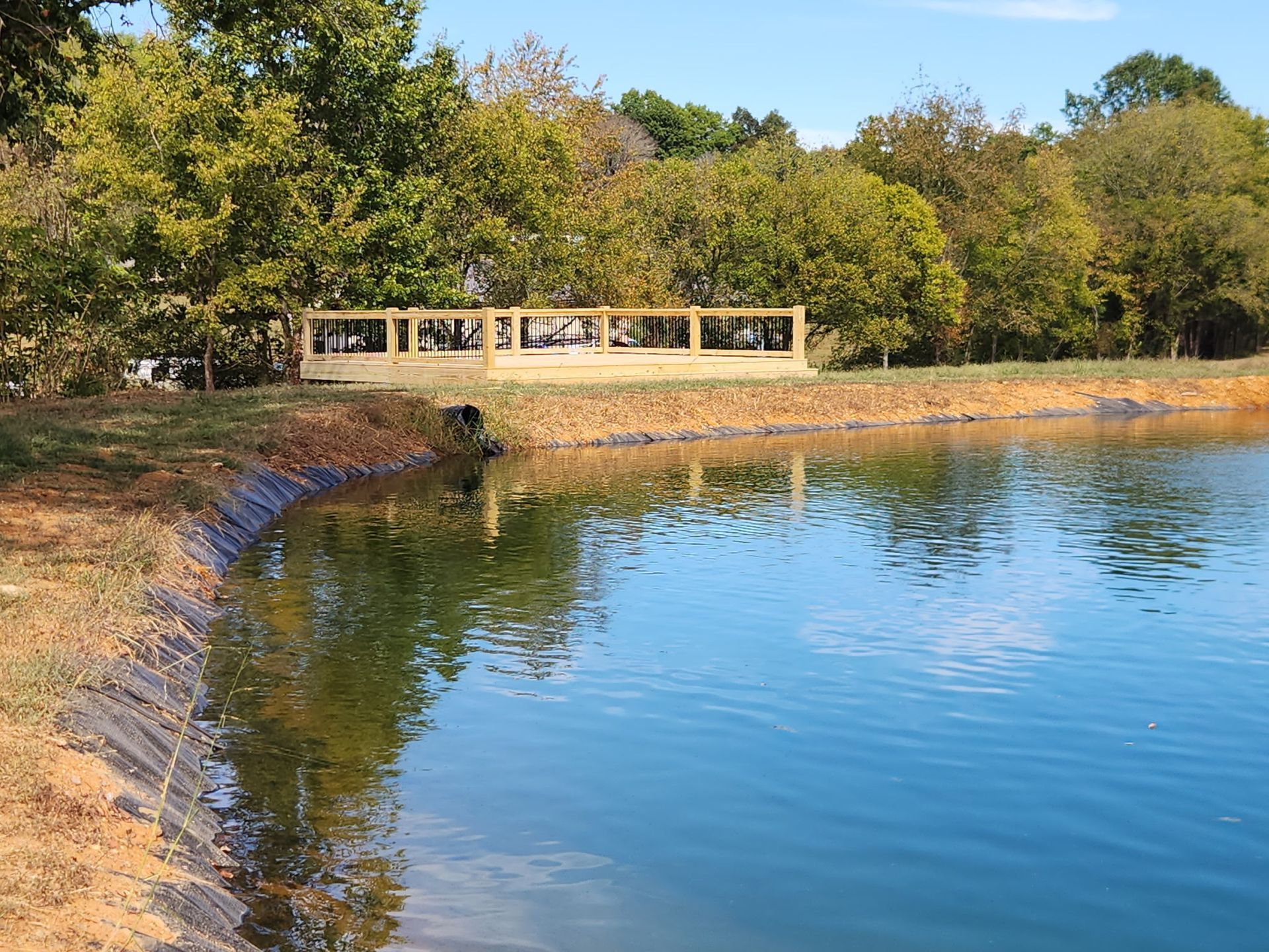 Calm lake with a curving rocky shore, reflecting trees and a small pavilion under autumn foliage.
