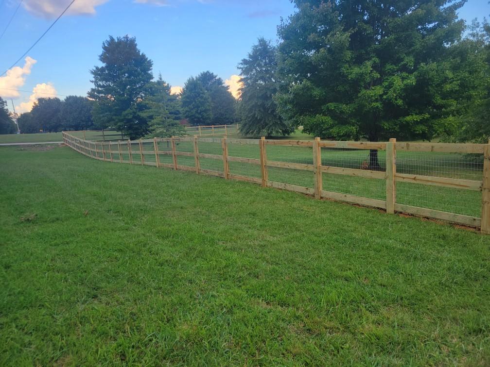 A wooden fence surrounds a grassy field with trees in the background.