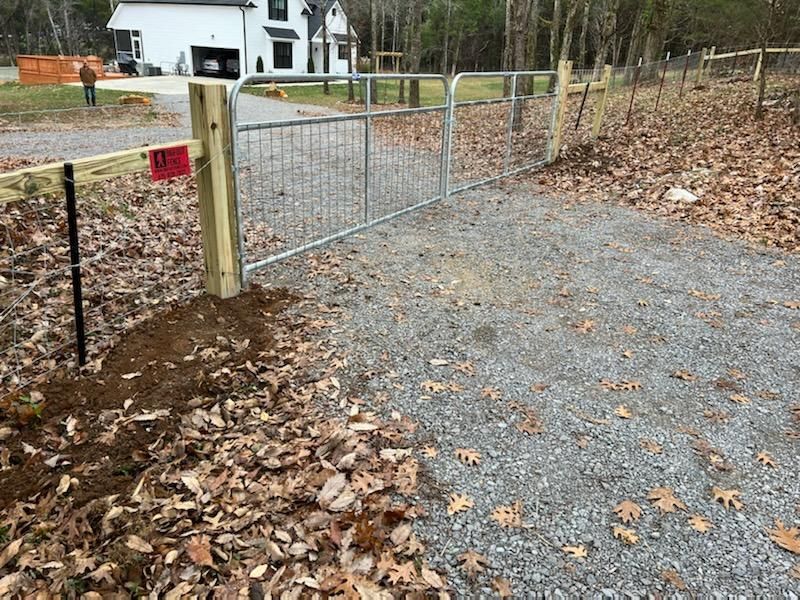 A metal gate is sitting on the side of a gravel road next to a wooden post.