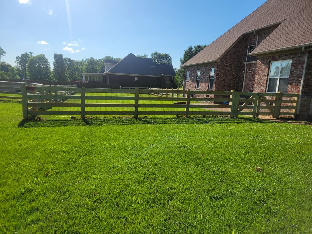 A wooden fence surrounds a lush green field in front of a house.