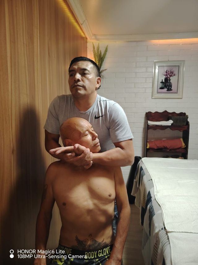Dos personas en un espacio cerrado; una está de pie detrás de la otra, con las manos alrededor de su cuello, en una habitación pequeña con una cama.