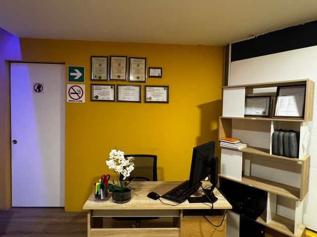 Pequeña zona de recepción de oficina con pared amarilla, escritorio, ordenador, silla y placas en la pared