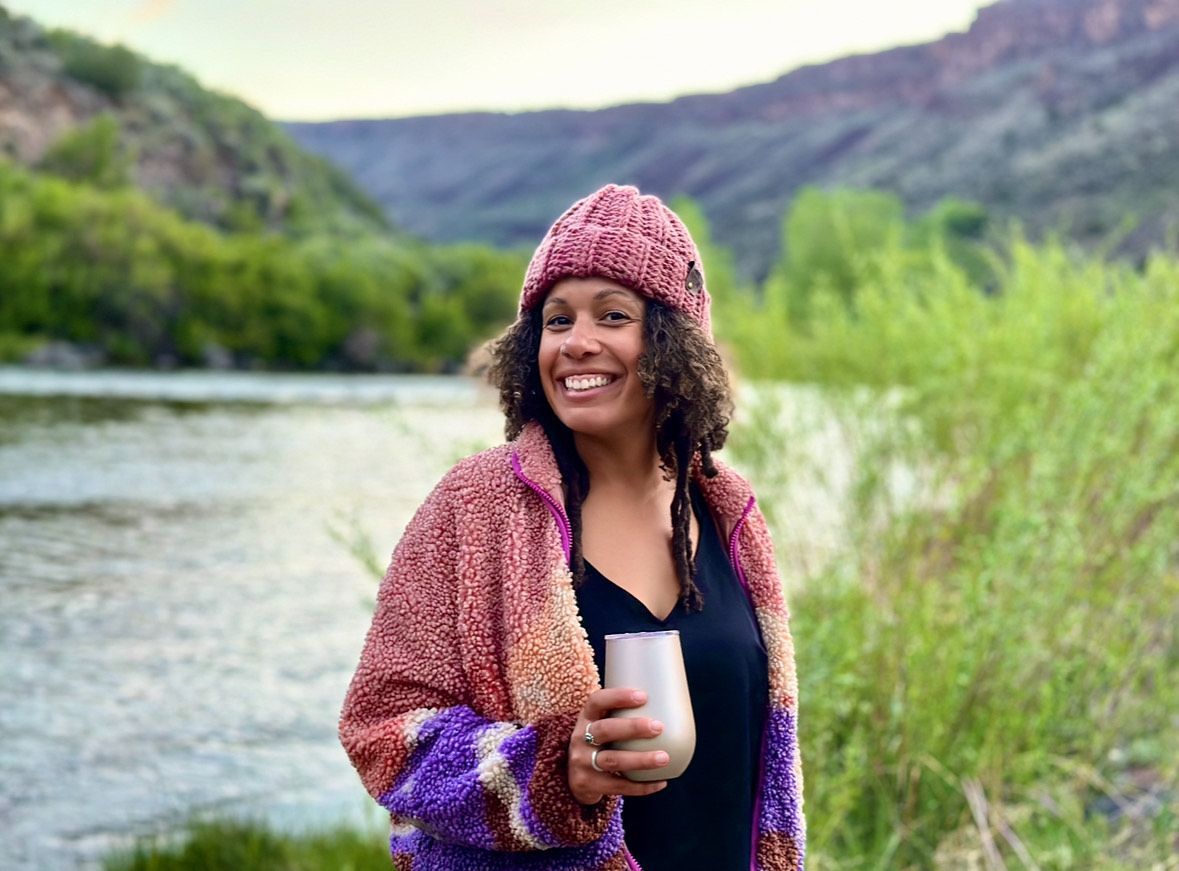 A woman is standing next to a river holding a glass of wine.
