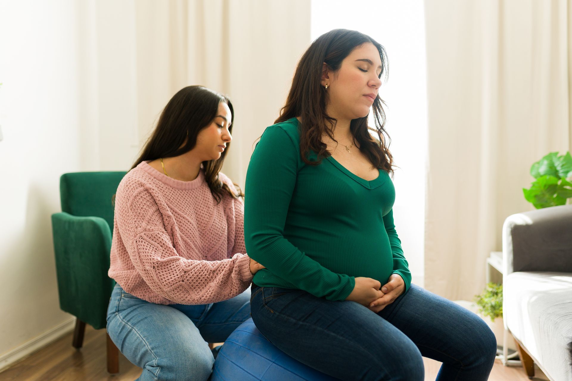 A person in a pink sweater provides support to another person sitting on a blue exercise ball in a brightly lit room.