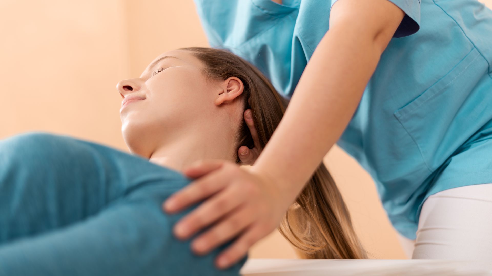 A practitioner in a teal uniform gently supports the neck and shoulders of a patient lying on a treatment table.