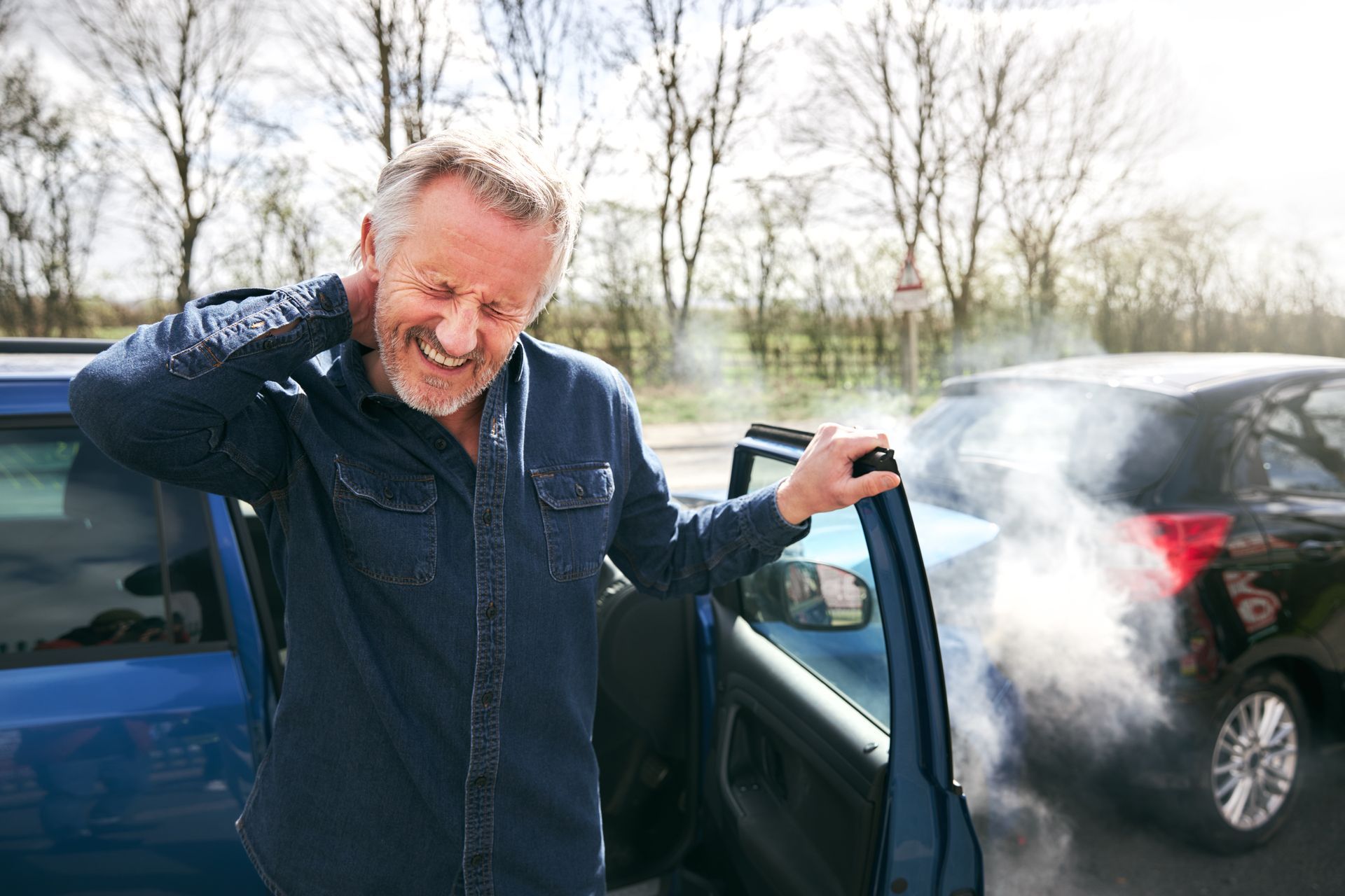 Person holding neck beside a damaged car with smoke rising after a traffic collision.