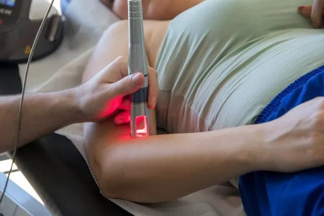 A hand holds a medical laser device emitting a red light against the skin of a person’s upper arm during treatment.