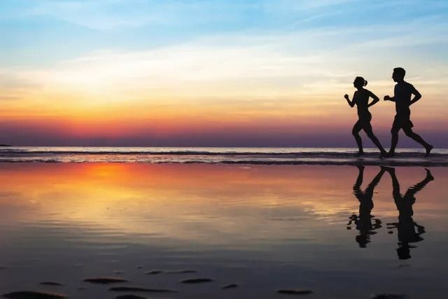 Silhouettes of two people jogging on a beach at sunset, with their reflections visible in the wet sand.