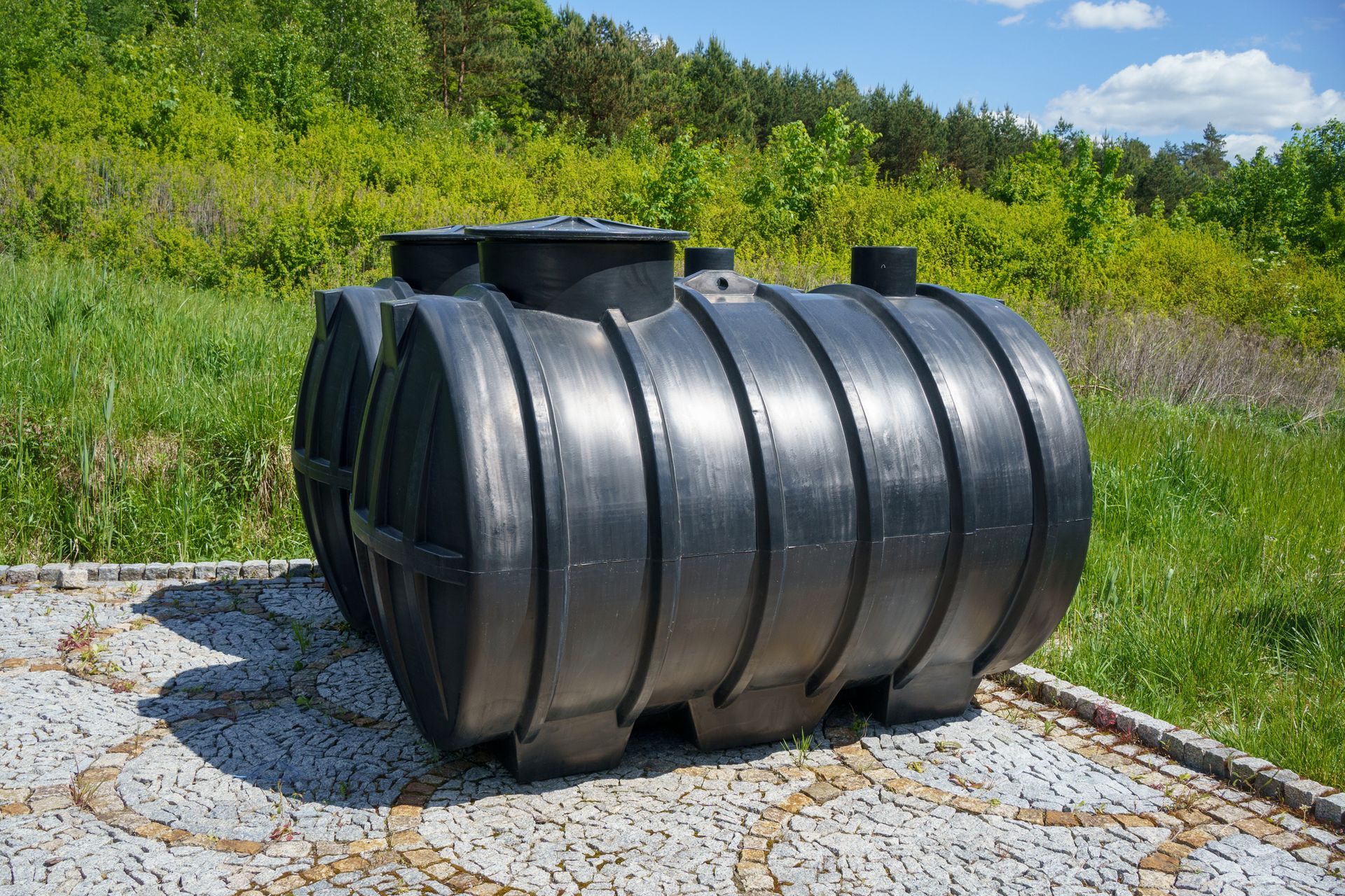 A large black plastic tank is sitting on a cobblestone driveway in a field.