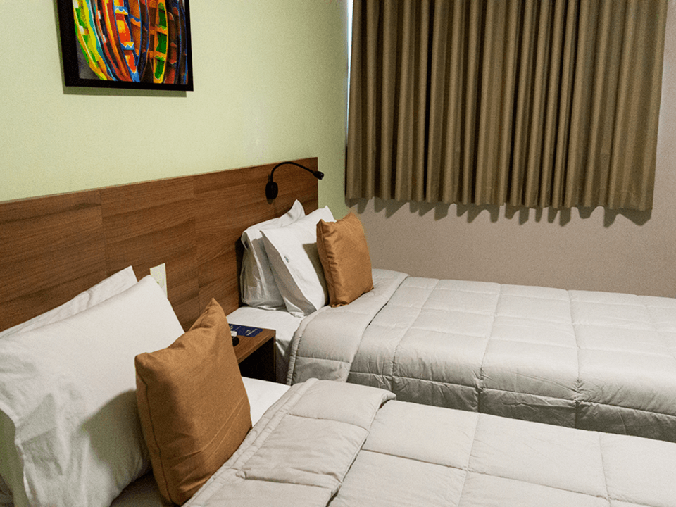 Duas camas de solteiro arrumadas com edredons brancos e almofadas decorativas em tom bege, posicionadas contra uma cabeceira de madeira em um quarto de hotel.