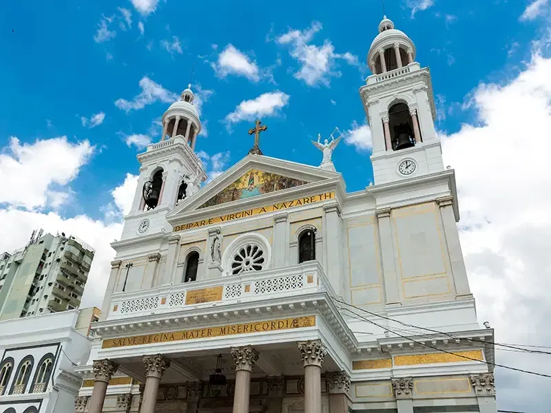 Uma igreja branca com duas torres sineiras, uma cruz central e inscrições em letras douradas contra um céu azul e nublado.