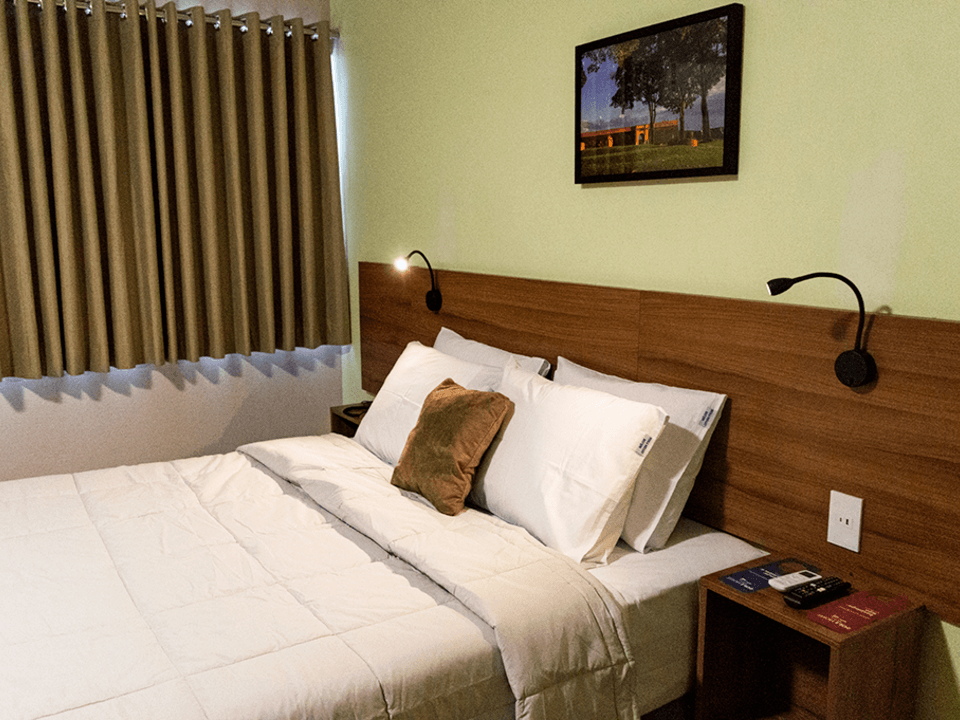 Uma cama arrumada com lençóis e travesseiros brancos, incluindo uma pequena almofada marrom, em um quarto com cabeceira e cortinas de madeira.