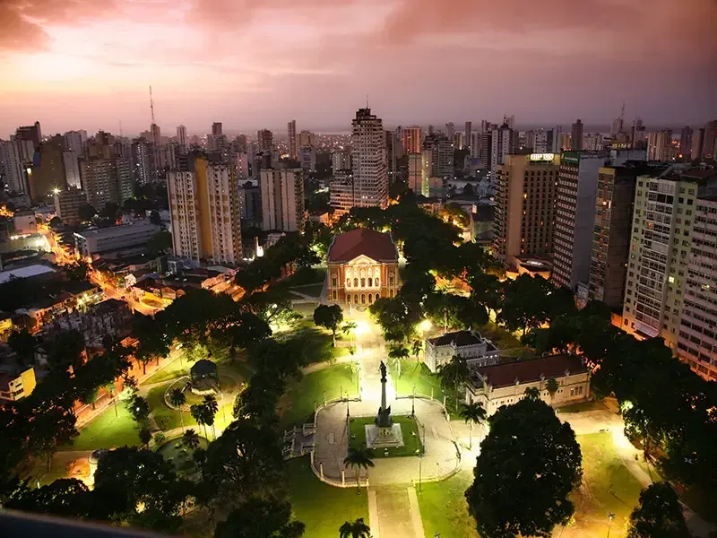 Vista aérea de um parque urbano ao entardecer, com um grande edifício, um monumento central e prédios altos ao redor.