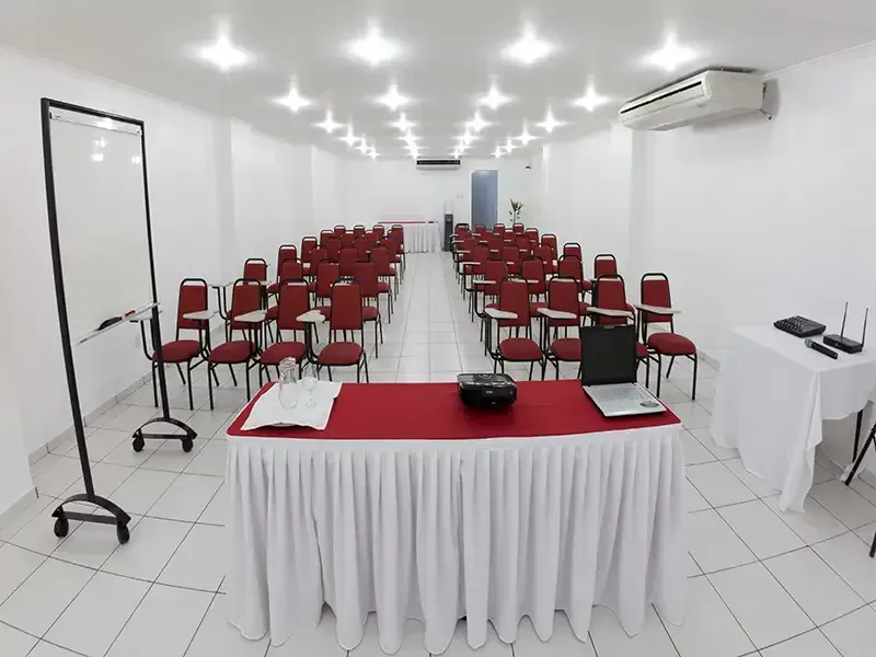 Uma sala de conferências ampla e iluminada, com fileiras de cadeiras voltadas para uma mesa de apresentações com um laptop, um projetor e um quadro branco.