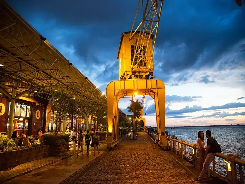 Um calçadão de paralelepípedos à beira-mar ao entardecer, com um grande guindaste industrial amarelo, prédios iluminados e pessoas.