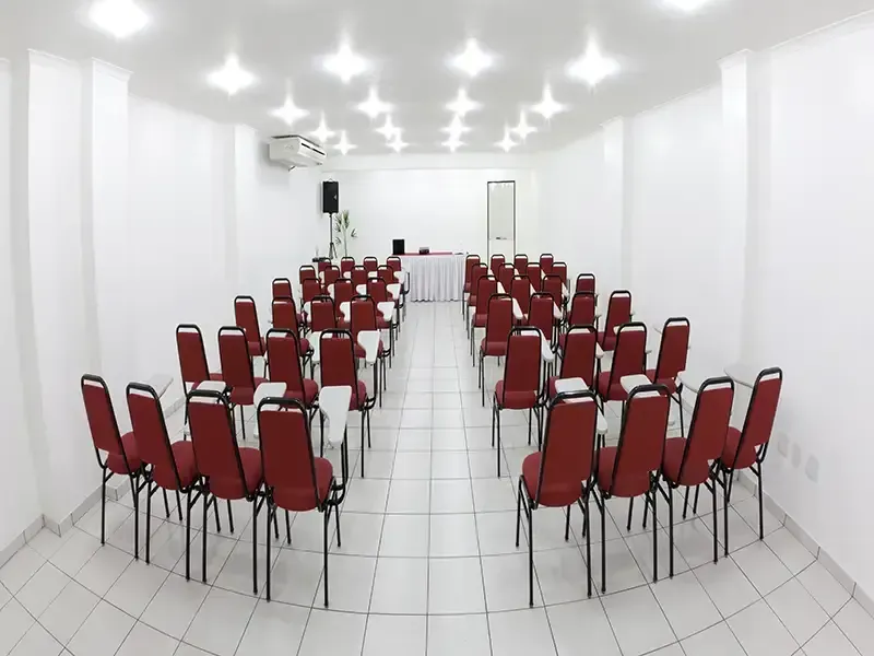 Uma sala de conferências ampla e iluminada, com fileiras de cadeiras cor de vinho dispostas em frente a uma pequena mesa, sobre um piso de azulejos brancos.