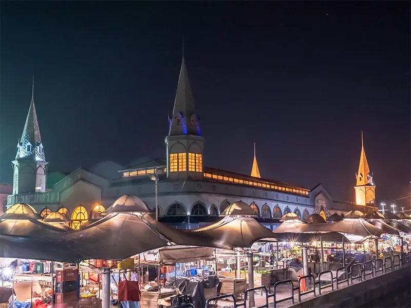 Barracas de mercado noturno montadas sob um edifício histórico com pináculos iluminados.
