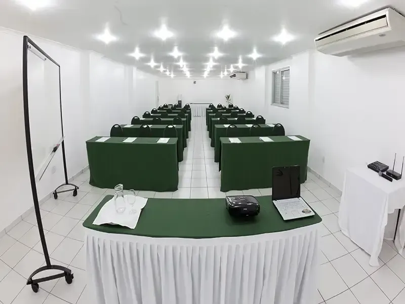 Uma sala de conferências com paredes brancas e fileiras de mesas cobertas com toalhas verdes, de frente para um pódio branco com um laptop.