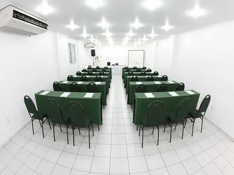Uma sala de conferências estéril e branca, com duas fileiras de mesas e cadeiras cobertas de verde, voltadas para um pódio e uma tela na frente.