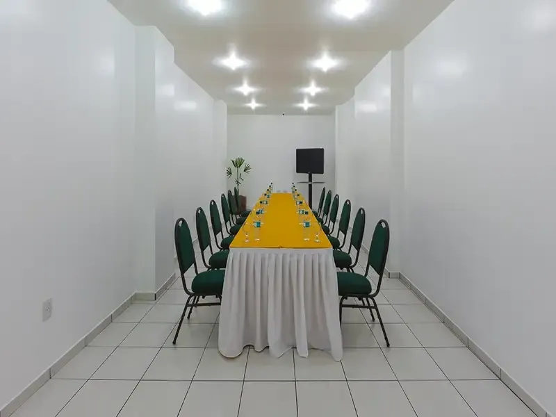 Uma longa mesa de conferência com uma toalha de mesa amarela e cadeiras verdes em uma sala iluminada com piso de azulejo e paredes brancas.