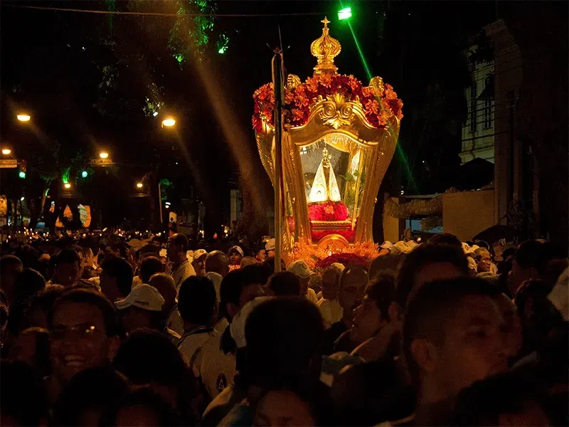 Uma multidão cerca um relicário religioso brilhante e ornamentado em ouro, que é carregado por uma rua à noite.