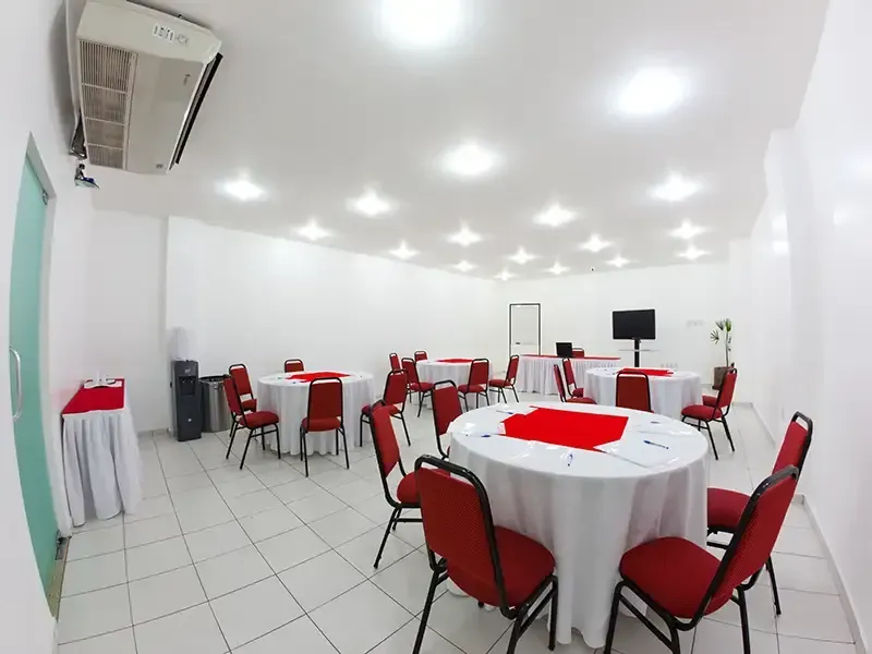 Uma sala de reuniões bem iluminada com várias mesas redondas cobertas com toalhas brancas, detalhes em vermelho e cadeiras vermelhas.
