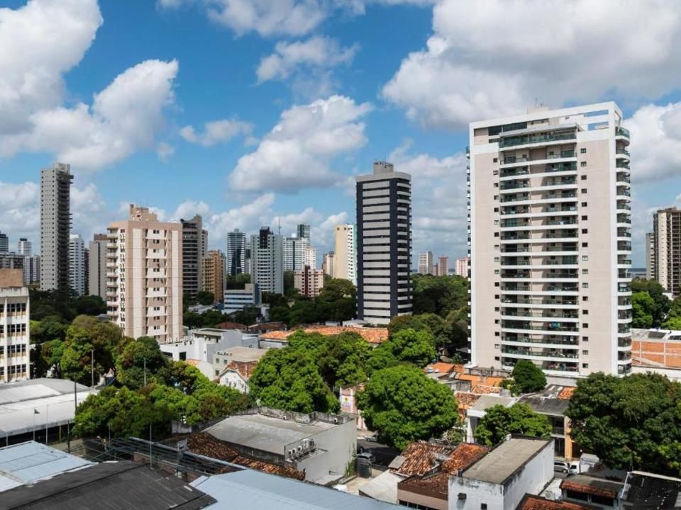 Vista aérea da linha do horizonte de uma cidade, com vários prédios altos de apartamentos rodeados por árvores exuberantes sob um céu azul.