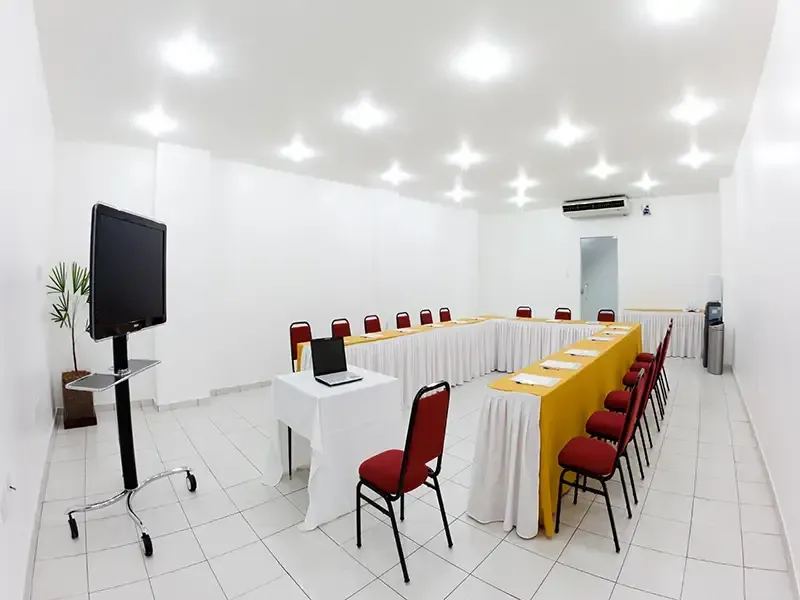Mesa de conferência em formato de U com cadeiras vermelhas em uma sala de reuniões clara e branca, com um monitor em um suporte com rodinhas.