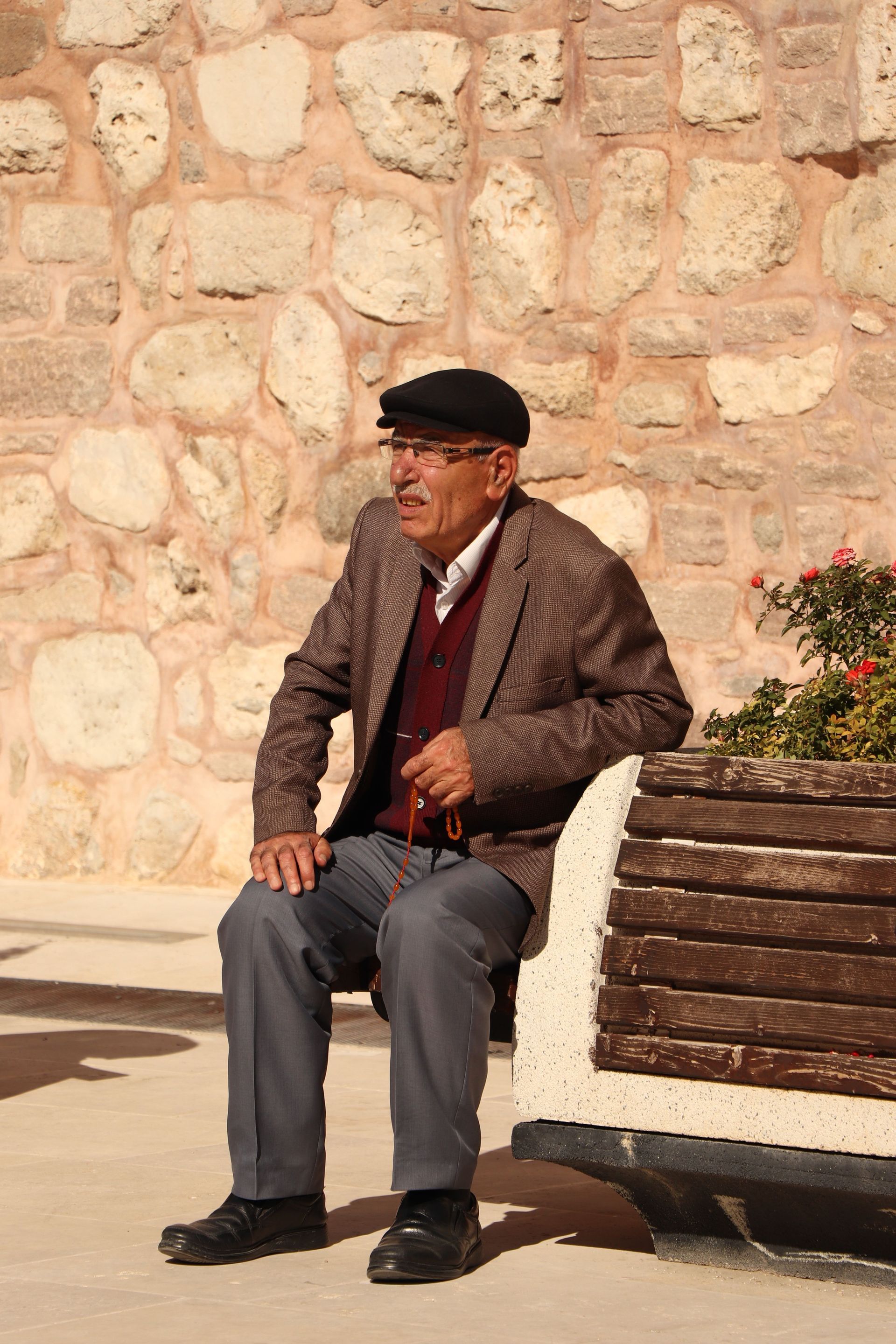 Man sitting on a bench outdoors against a stone wall, wearing a dark cap and brown jacket.