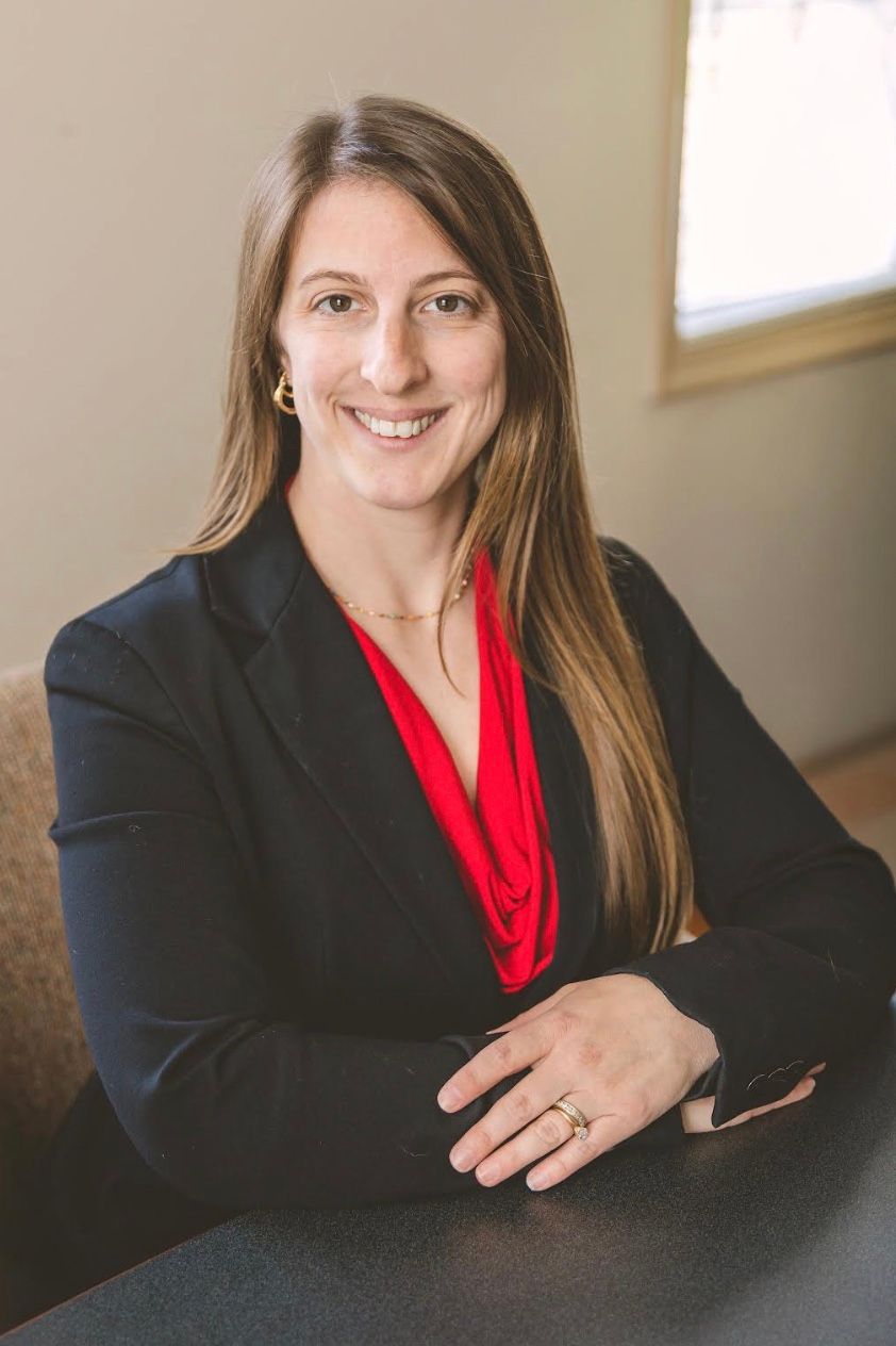 Profesional sonriente, vestida con un blazer negro y una blusa roja, sentada en un escritorio en una oficina.