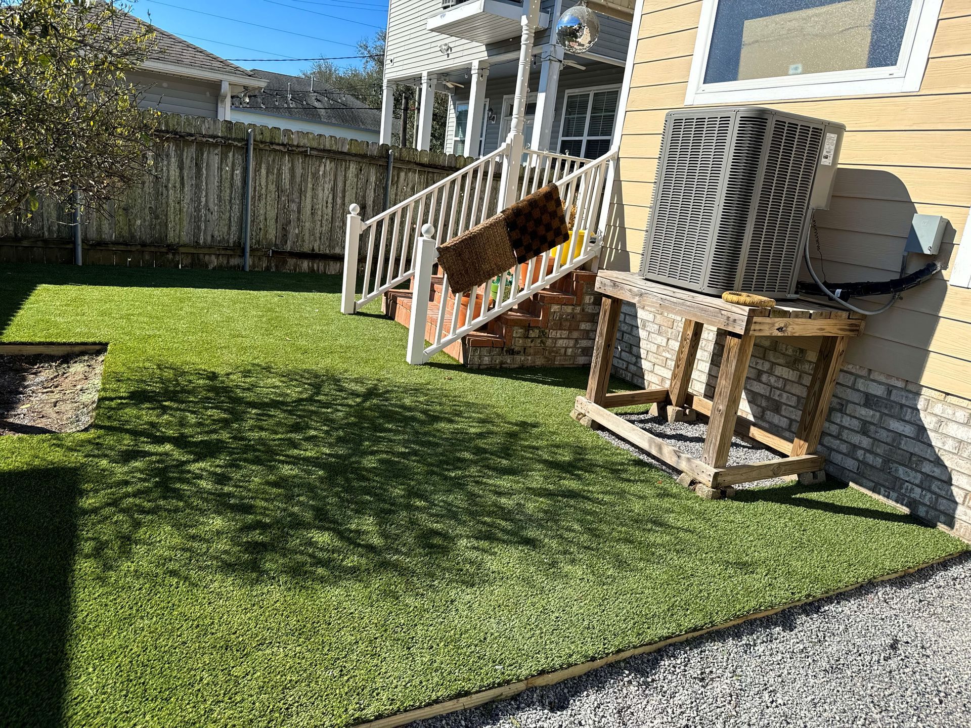 Backyard with green lawn, wooden stairs, white fence, and an air conditioner beside a yellow house.