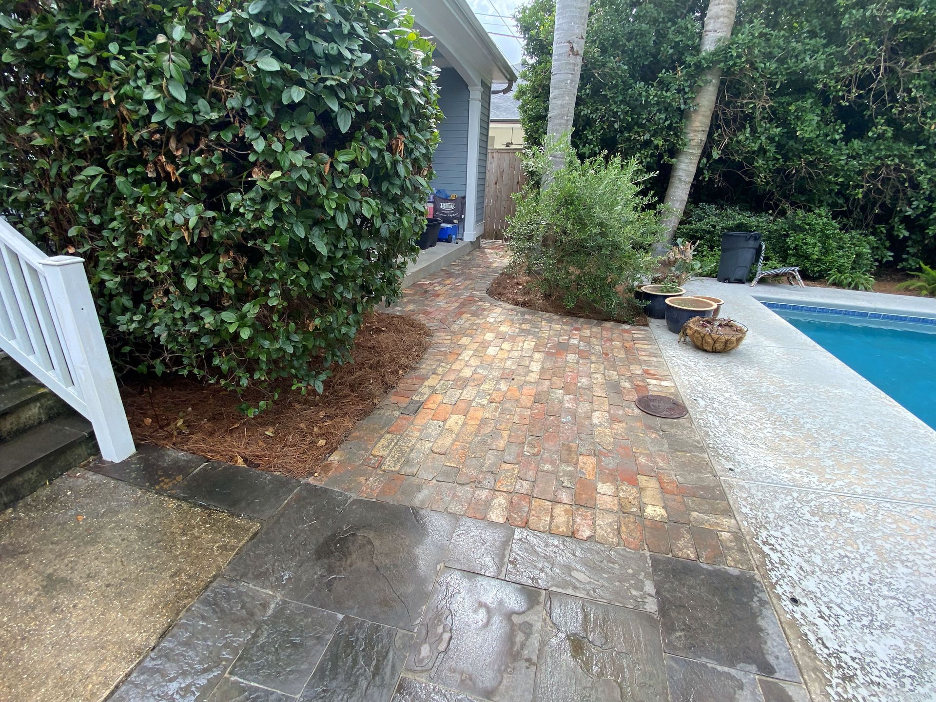 Brick patio beside a pool, with wet stone pavers, shrubs, and a white fence