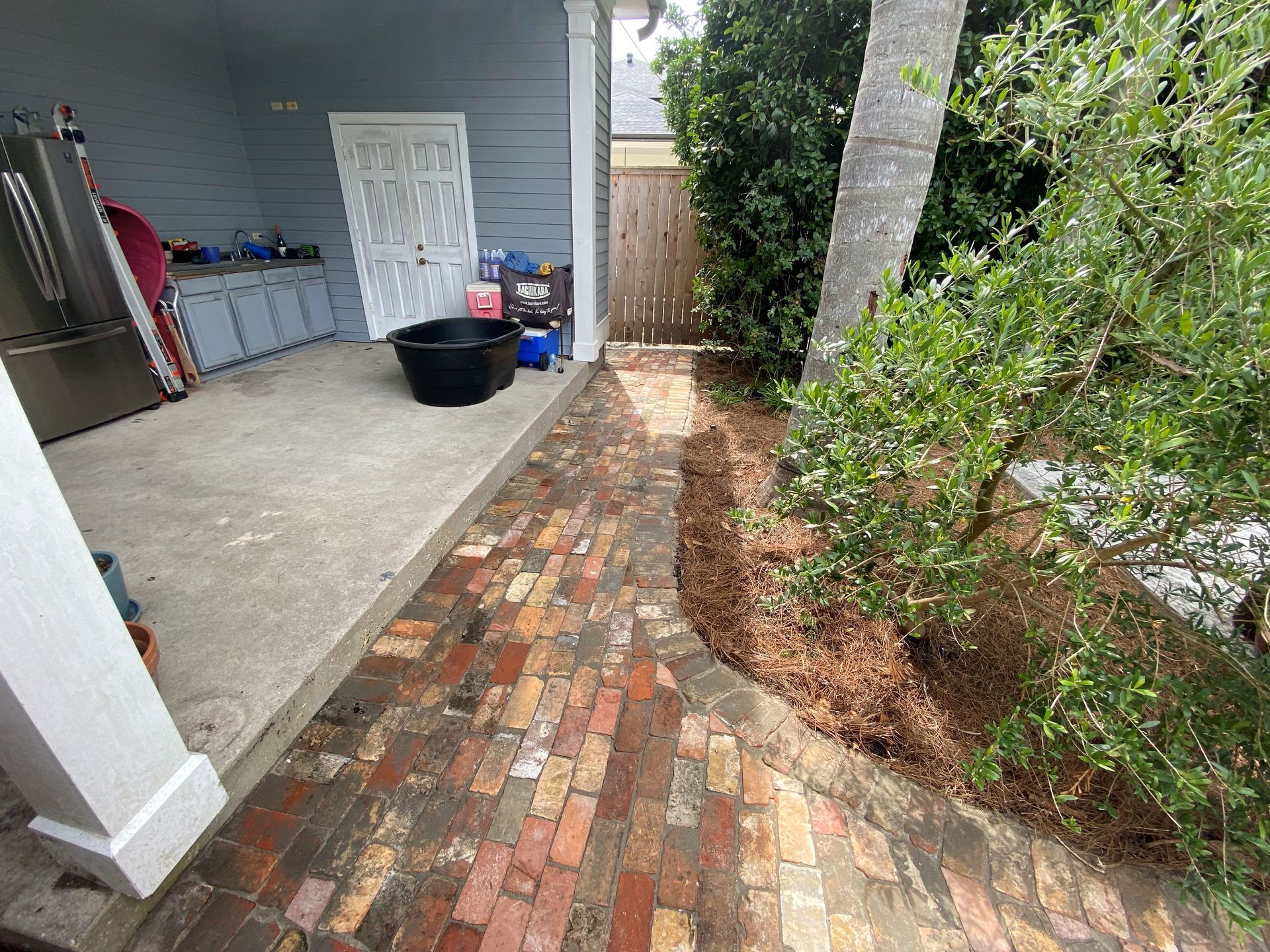 Brick walkway beside a gray house and porch, bordered by shrubs and a large tree.