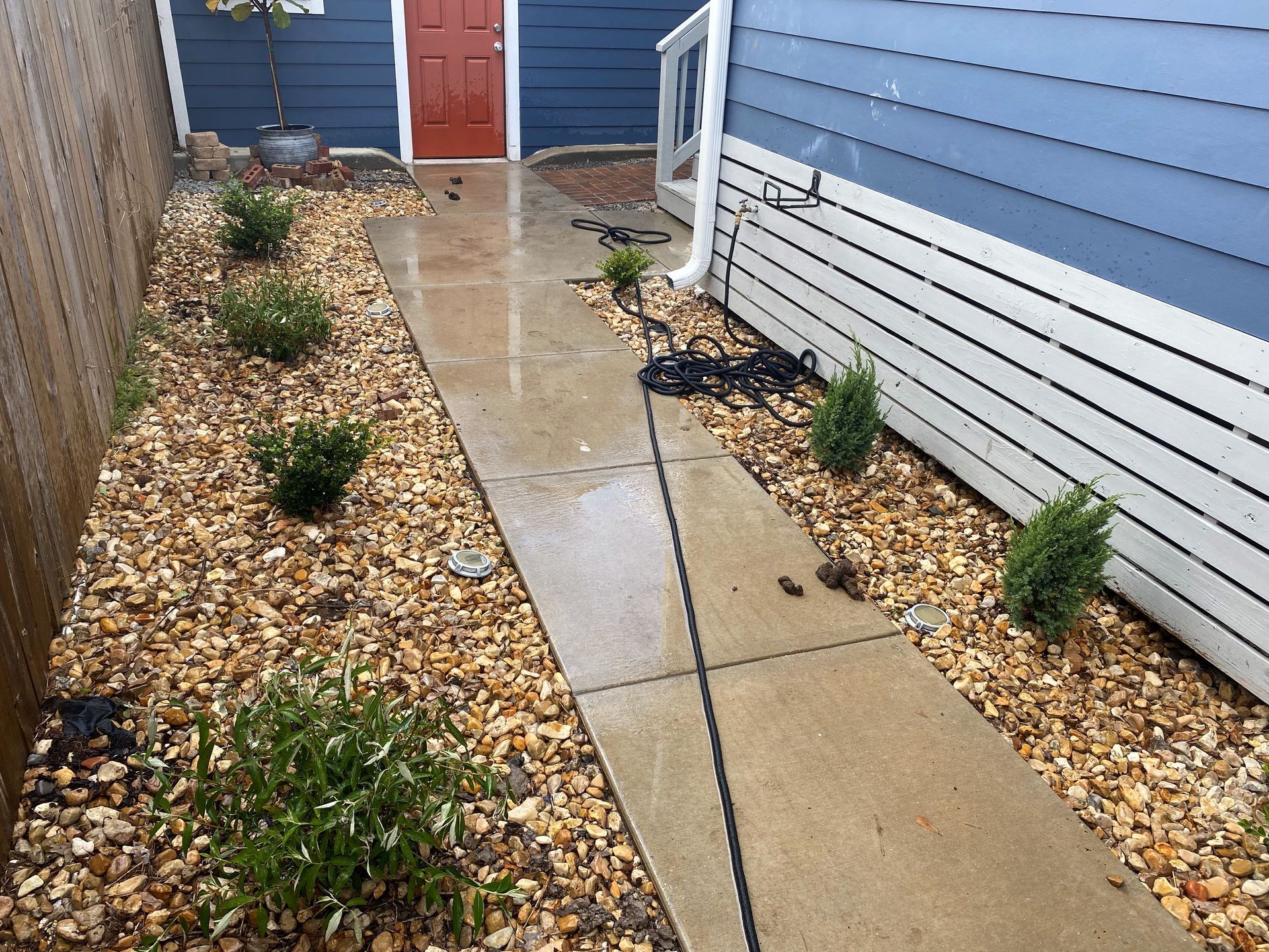 Wet concrete walkway between houses with gravel beds and a red door in the distance