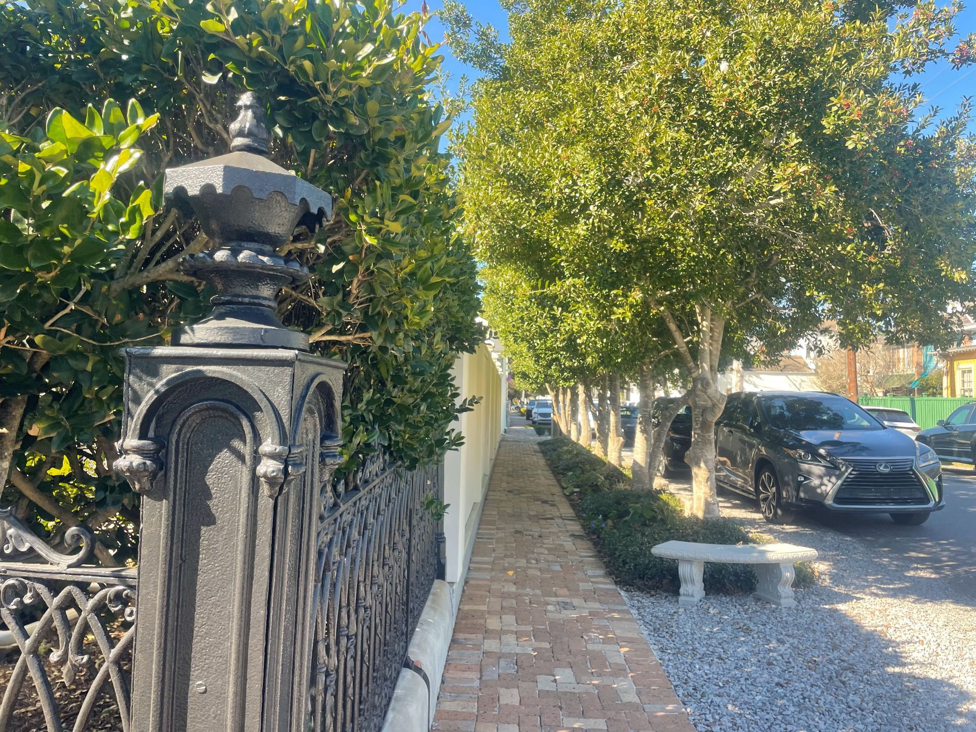 Sunlit sidewalk beside an ornate black fence and tree-lined gravel drive with parked cars