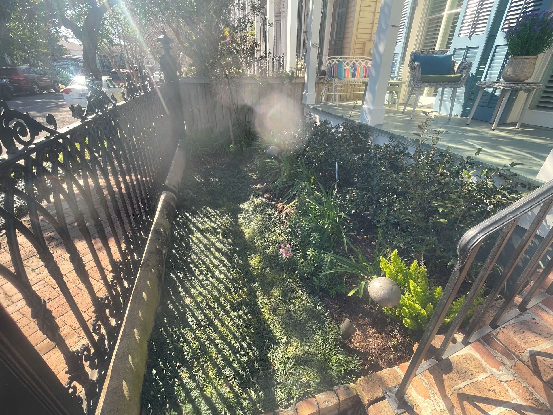 Black metal fence around a small landscaped garden beside a brick walkway in bright sunlight