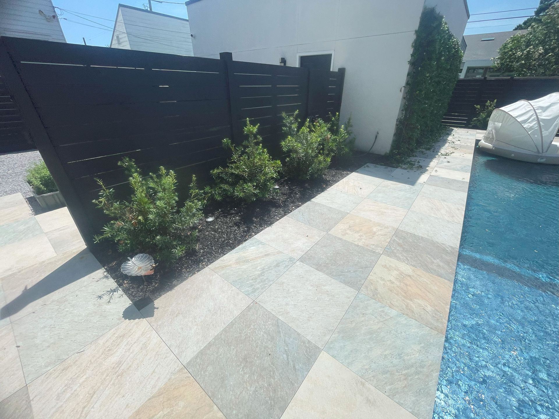 Patio with gray stone pavers, black privacy fence, shrubs, and a blue pool edge on the right
