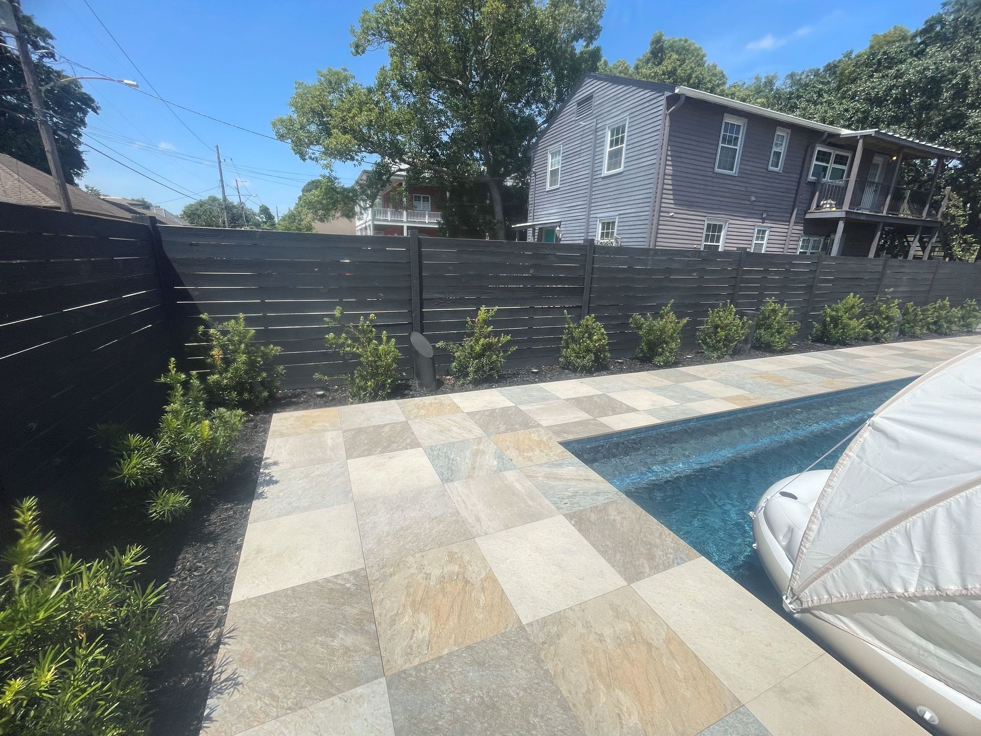 Backyard patio with pool, stone tiles, black fence, and trees under a blue sky