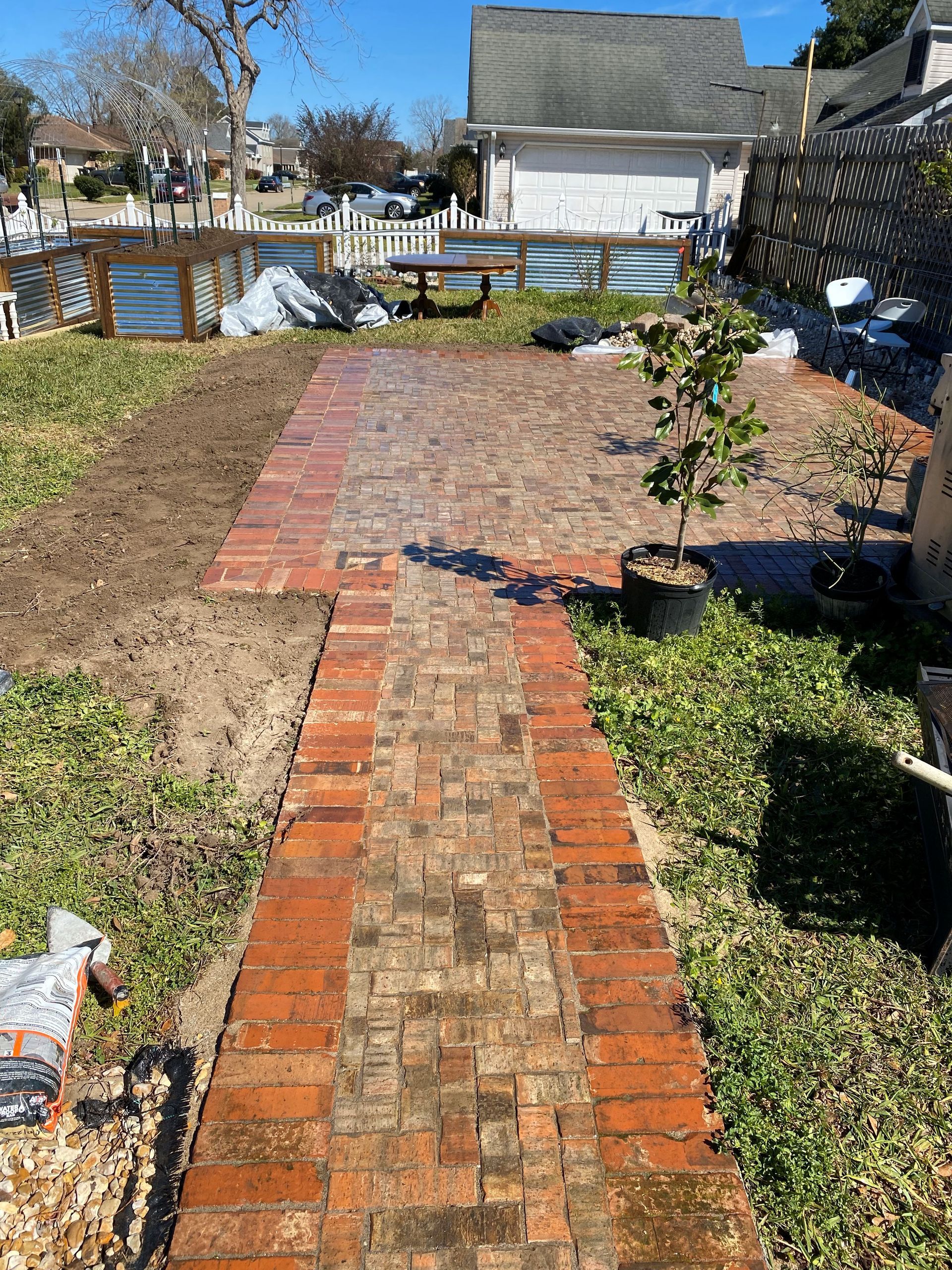 Brick garden path leading to a backyard patio, bordered by mulch and low plants under a sunny sky