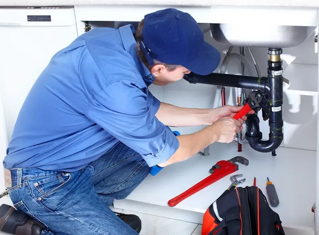 A plumber, wearing a blue cap and shirt, kneels under a kitchen sink, using a wrench to repair pipes.