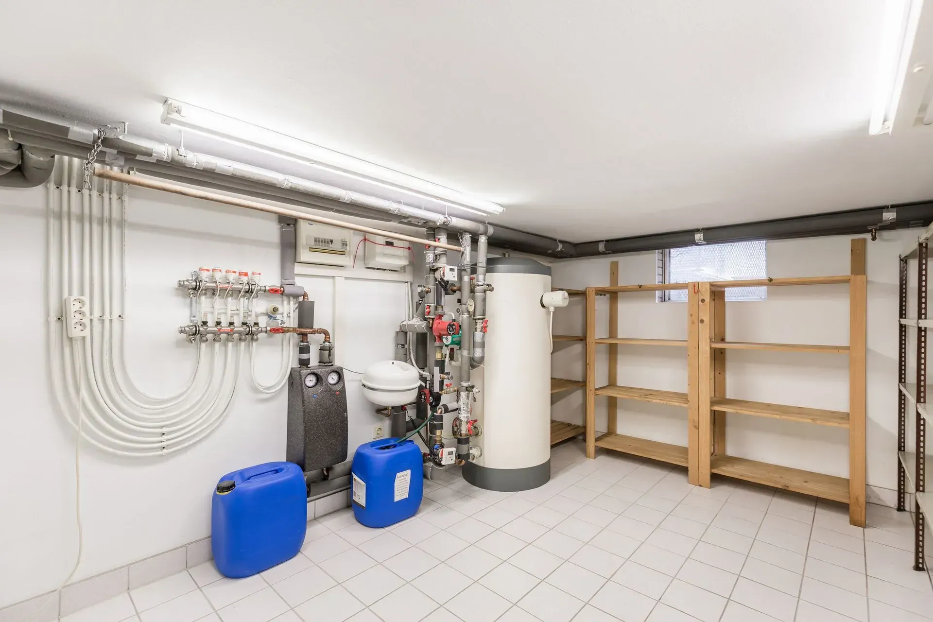A basement utility room with a water heater, piping, blue containers, and wooden shelves. The room has white walls and a tiled floor.