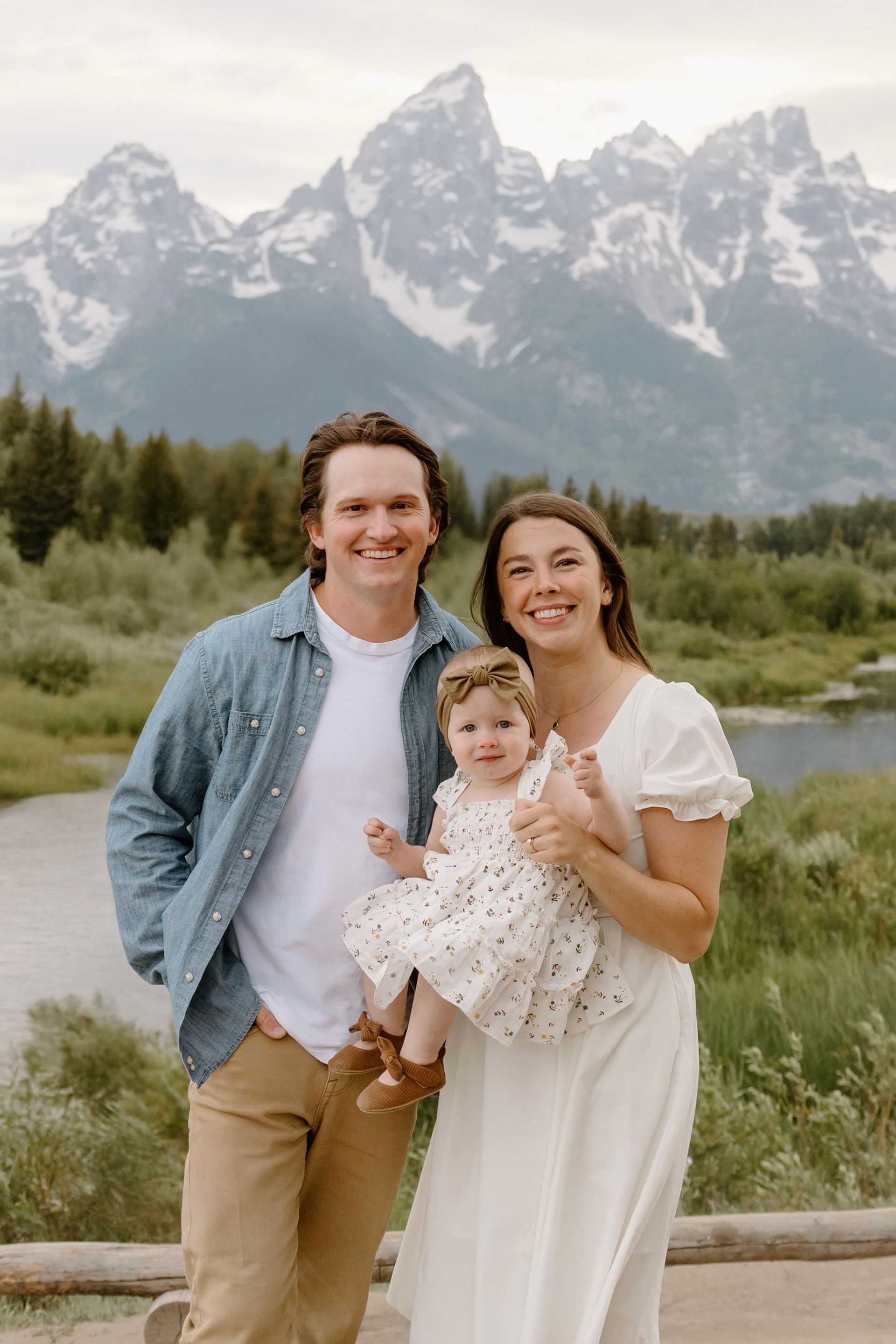 Family poses in front of mountains; man in denim shirt, woman in white dress, child in floral dress.