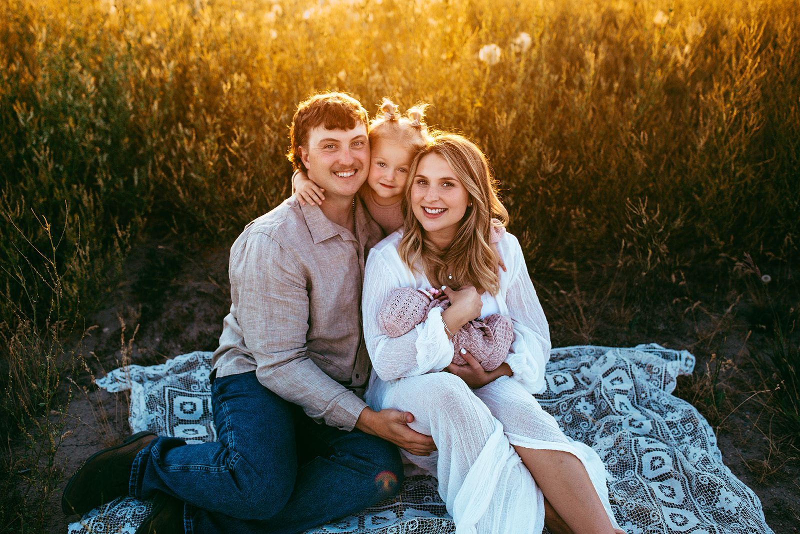 Family of four smiles for the camera while sitting on a blanket in a field at sunset.