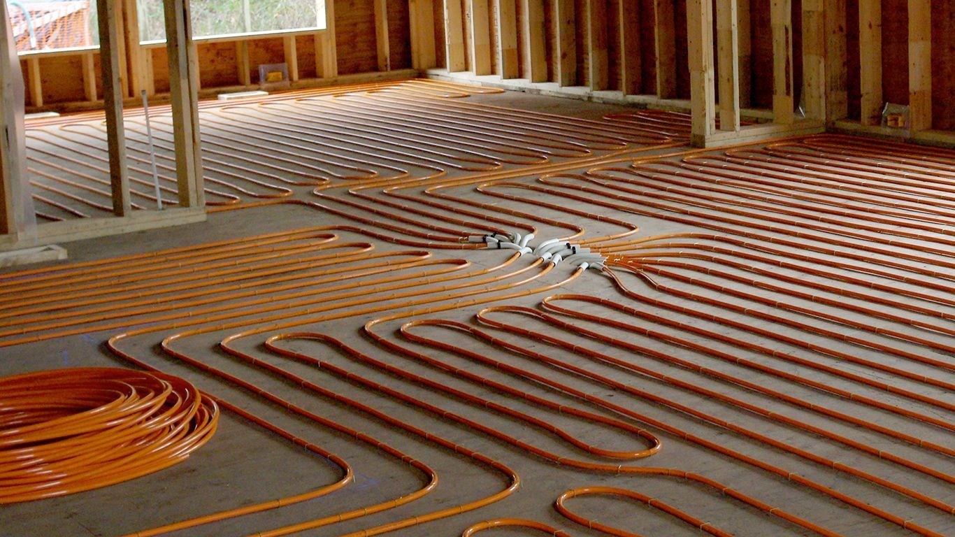Orange tubing laid across a concrete floor, likely for radiant floor heating in a building under construction. Wooden framing is visible.